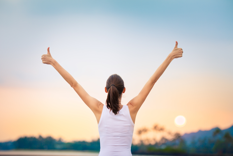 woman holding her arms up in the air giving thumbs up facing the sunset